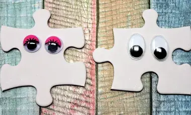 Two white jigsaw puzzle pieces side by side decorated with wiggle eyes against a backdrop of different colored stained wood.