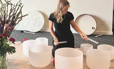 A woman sitting in the middle of six variously sized glass bowls, looking down with the playing sticks.