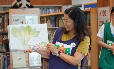 Smiling person wears a purple apron with the word STAR on it. The person is standing in front of library shelves and holding up a picture book.