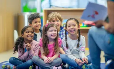 5 small smiling children sitting on a rug facing an adult holding a book. 