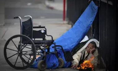 Person on the street huddled under tarp next to wheel chair with a fire burning