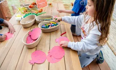 Three children sit at a wooden table with art supplies in small baskets, decorating pink paper fruits.