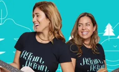 Two smiling women with guitars, wearing t-shirts that say Again Again