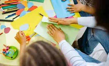 An assortment of craft supplies, paints, markers and hands with scissors cutting paper.