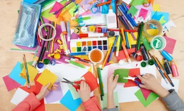 Children's hands holding scissors and cutting paper with a table full of arts and crafts supplies