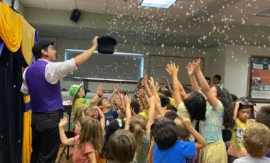 A man in a purple vest, standing in front of a yellow curtain, holding up a black top hat. There are bubbles coming out of the hat, and a crowd of young children are reaching up towards the bubbles.