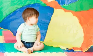 Baby sits on a colorful floor in front of an equally colorful parachute.