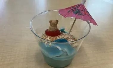 A cup of blue pudding on a table decorated with cookie crumbs, candy and a paper umbrella