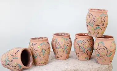 Six small, terracotta-colored ceramic cups, painted with teal and lime green swirling patterns, are arranged on a textured stone block against a white background. One cup is lying on its side.