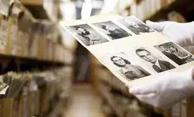 In the foreground are white gloved hands holding a white sheet with black-and-white photographs of people. In the background are rows of boxes on shelves.