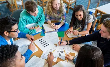 A group of seven teenage girls and boys seated around a round table with documents, notebooks and opened textbooks scattered on the surface.