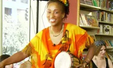 Ina in yellow and orange robes holding a jimbe drum.