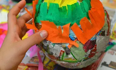A child’s hand applies cut tissue paper to a paper mache form.