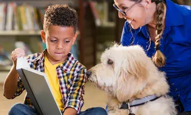 a young boy shows a book to an attentive dog and its adult handler