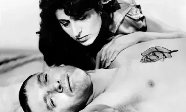 The male protagonist is on bed the female protagonist is bent over him.