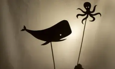 Two hands holding up a shadow puppet of a whale and squid on sticks between a light source and a white fabric to make shadows appear.