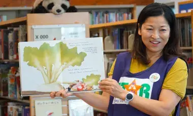 A woman woman wearing a STAR volunteer vest holds up a picture book in a library