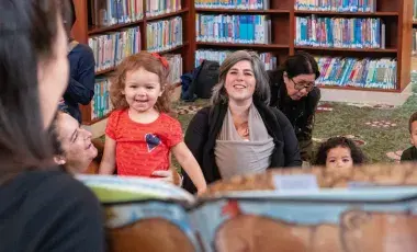 Several children and adults watch a woman with long dark hair holding a picture book.