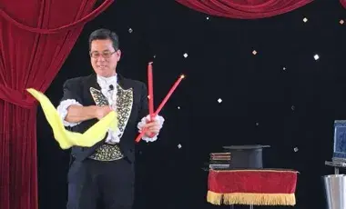 A magician holding wands and a scarf on stage next to a table with a top hat and a cage