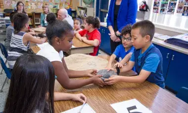 Three children gathered around a table touch an artifact at the center of the table. Additional children seated at a table nearby look at a similar artifact.