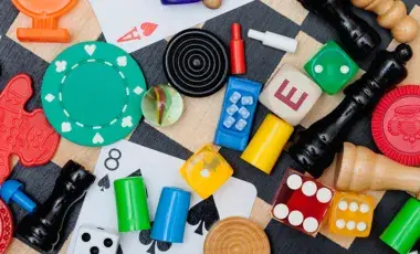 Assorted board game pieces, poker chips, dice, chess pieces, playing cards, and letter blocks scattered on a checkered surface.