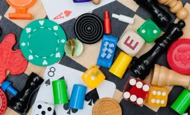 Colorful board-game pieces, cards, dice, chips, pawns, and wooden tokens on a tabletop scene on a checkered background.
