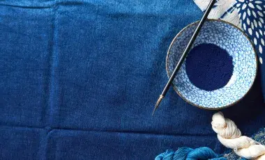 Fabric in various shades of blue topped with a patterned bowl containing blue powder and paint brush and blue and white fabric along the top and bottom.