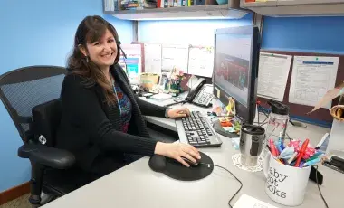 A librarian sits at a desk in an office. She is wearing a phone headset and smiling at the viewer. 