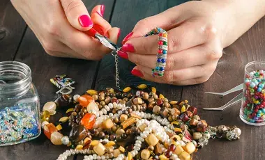 A pair of hands makes a piece of jewelry with pliers and assorted beads.