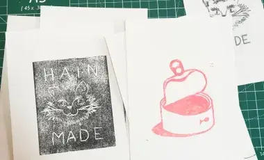 Example linocut prints on cards, one with a cat face that says "Hain Made" and another with an open can of food.