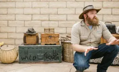 A bearded adult man crouched in front of a block wall with various baskets and trunks behind him.