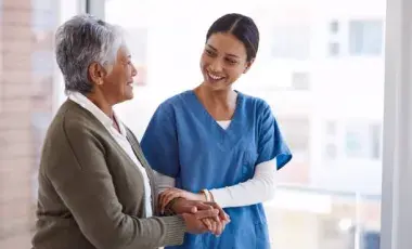 older adult dressed in a brown sweater walking with nurse