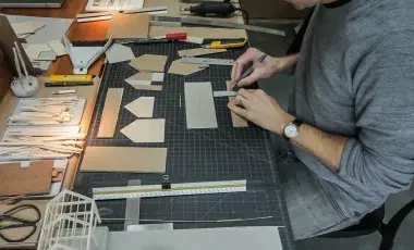 Person measuring and cutting cardboard pieces on a gridded work table with various crafting tools and materials around.