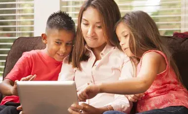 a mother holds a computer tablet in her lap, sharing it with a young boy and girl sitting on either side