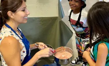 An instructor in an apron showing two children how to stir food in a pot.