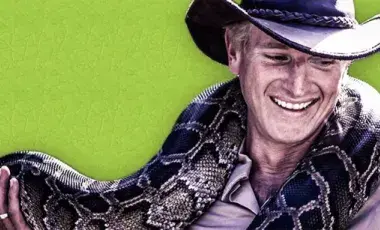 A man wearing a cowboy hat is smiling while holding onto a brown spotted python. 
