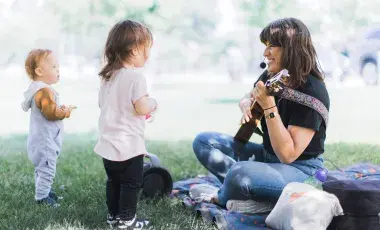 Leah is seated cross-legged on a grassy lawn holding her guitar. She is mid-song and smiling at two toddlers. 