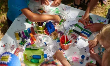 Children crafting with beads, and glue at a table covered with art supplies.