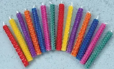 Fourteen small, multi-colored rolled beeswax candles are arranged in an arc on a plain gray background. The candles are unlit.