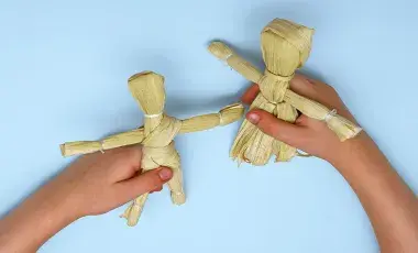 Hands hold two human shaped dolls made out of corn husks