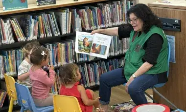 Ms. Judy reads aloud to three children. She is seated in front of a shelf of picture books and is wearing a green STAR volunteer vest.