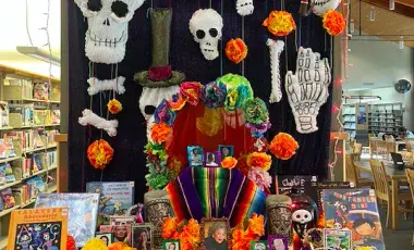 Community altar/ofrenda for Día de Muertos displayed in a library. Decorated with orange flowers, paper skulls, puffy fabric skulls and books