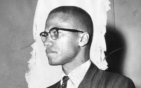 Black and white photo of Malcolm X from the side, standing at a meeting.