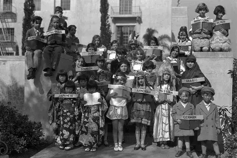 Children dressed in the costumes of many different countries hold signs indicating the languages in which books are available at the Los Angeles Public Library, ca 1939 