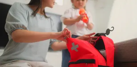 A woman packs a red backpack with a medical cross on it while a child holds a water bottle