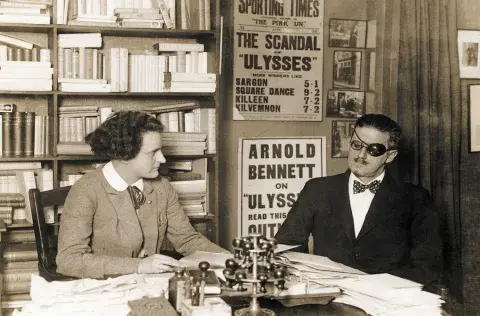 James Joyce with Sylvia Beach sitting next to each other at a desk.