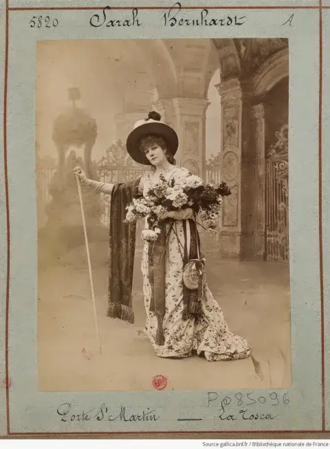 Sarah in an long dress and hat, holding flowers and a staff.