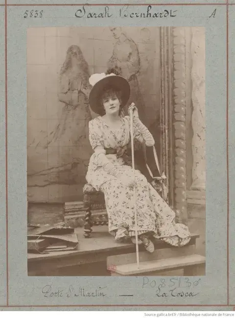 Sarah sitting down wearing a long dress, hat and with a staff on hand.