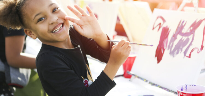 A young girl waving at the camera while holding a paint brush in her right hand