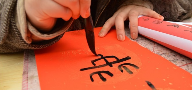 Hand with brush writing Chinese calligraphy on red paper.